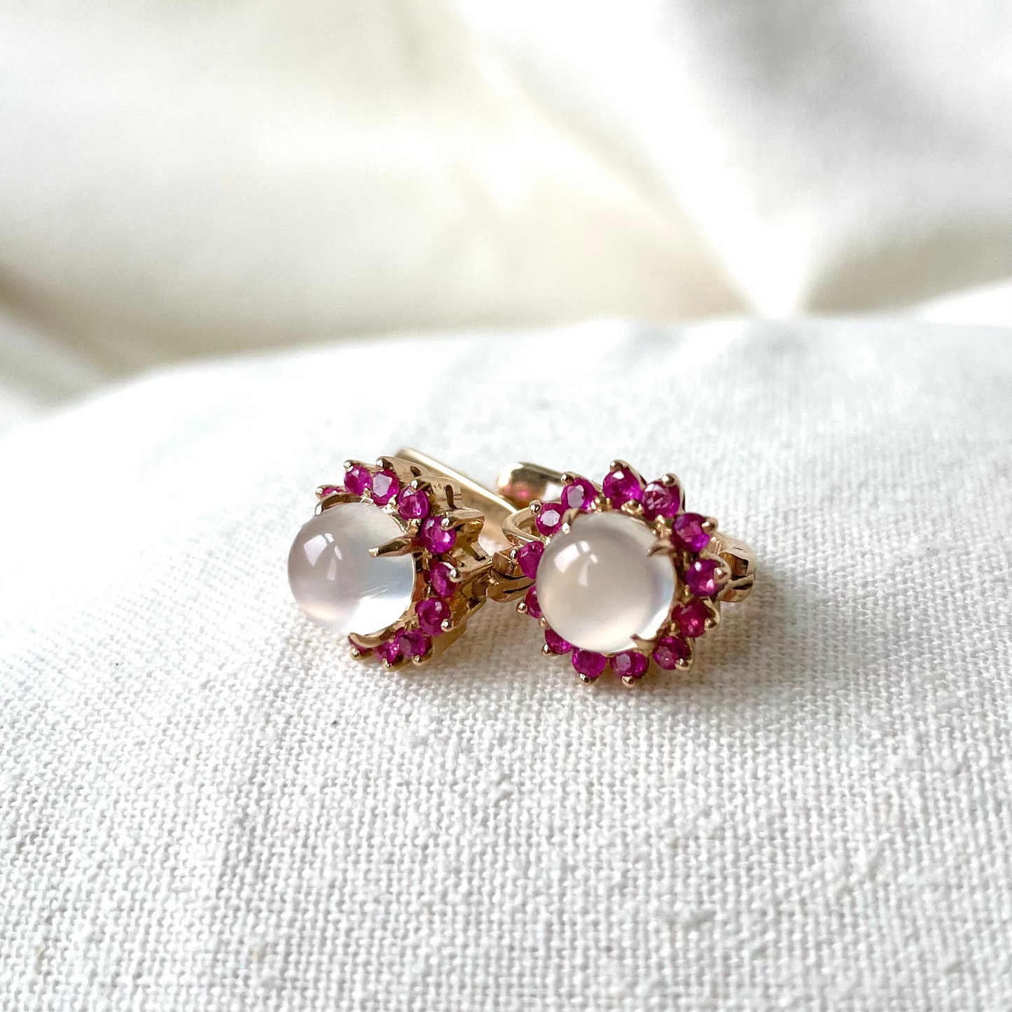 A pair of 14 K yellow gold lever back earrings with moonstone centers and ruby accents, placed on a white fabric surface.