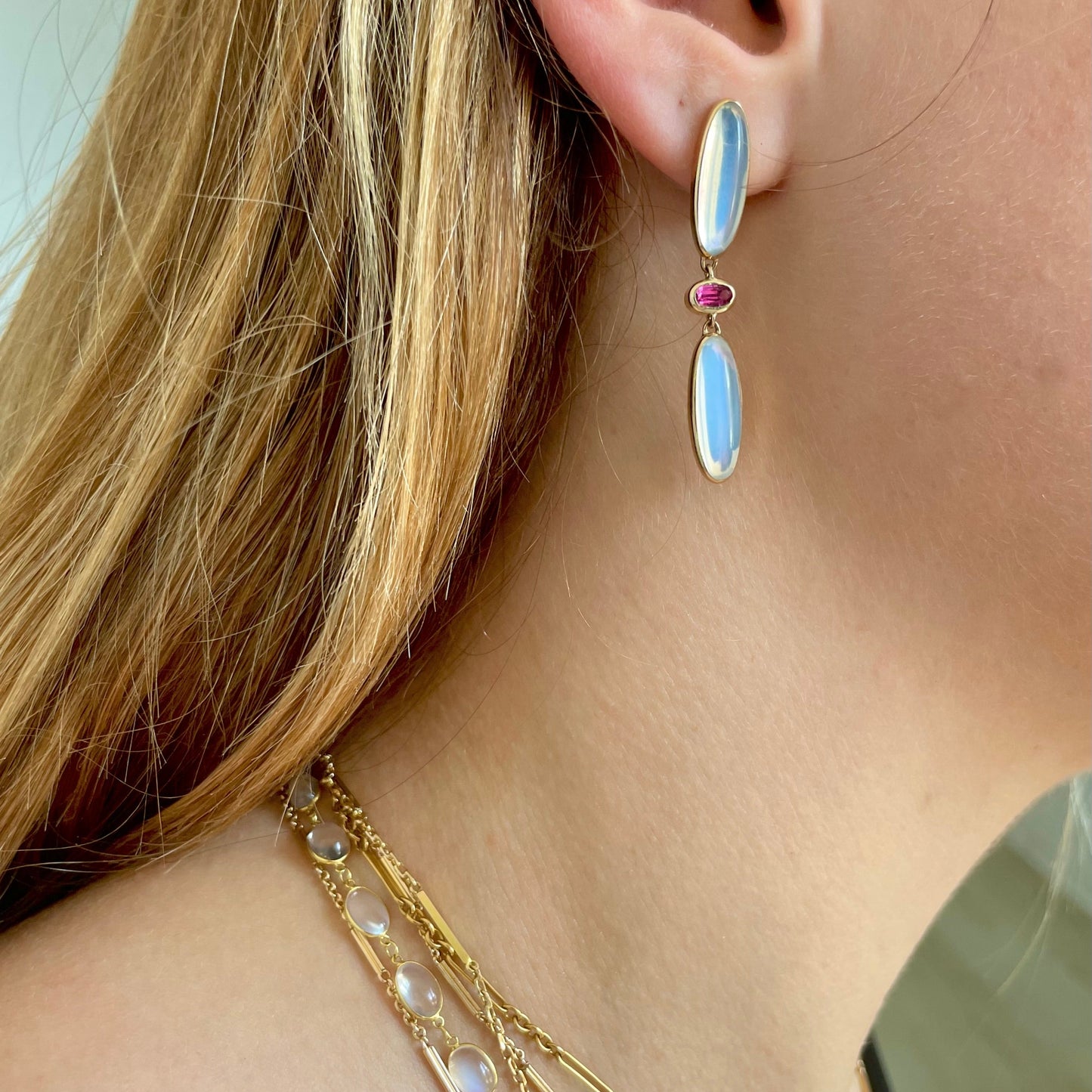 Close-up of a model wearing a gold earring with a pink tourmaline and moonstone, with and multiple gold necklaces.