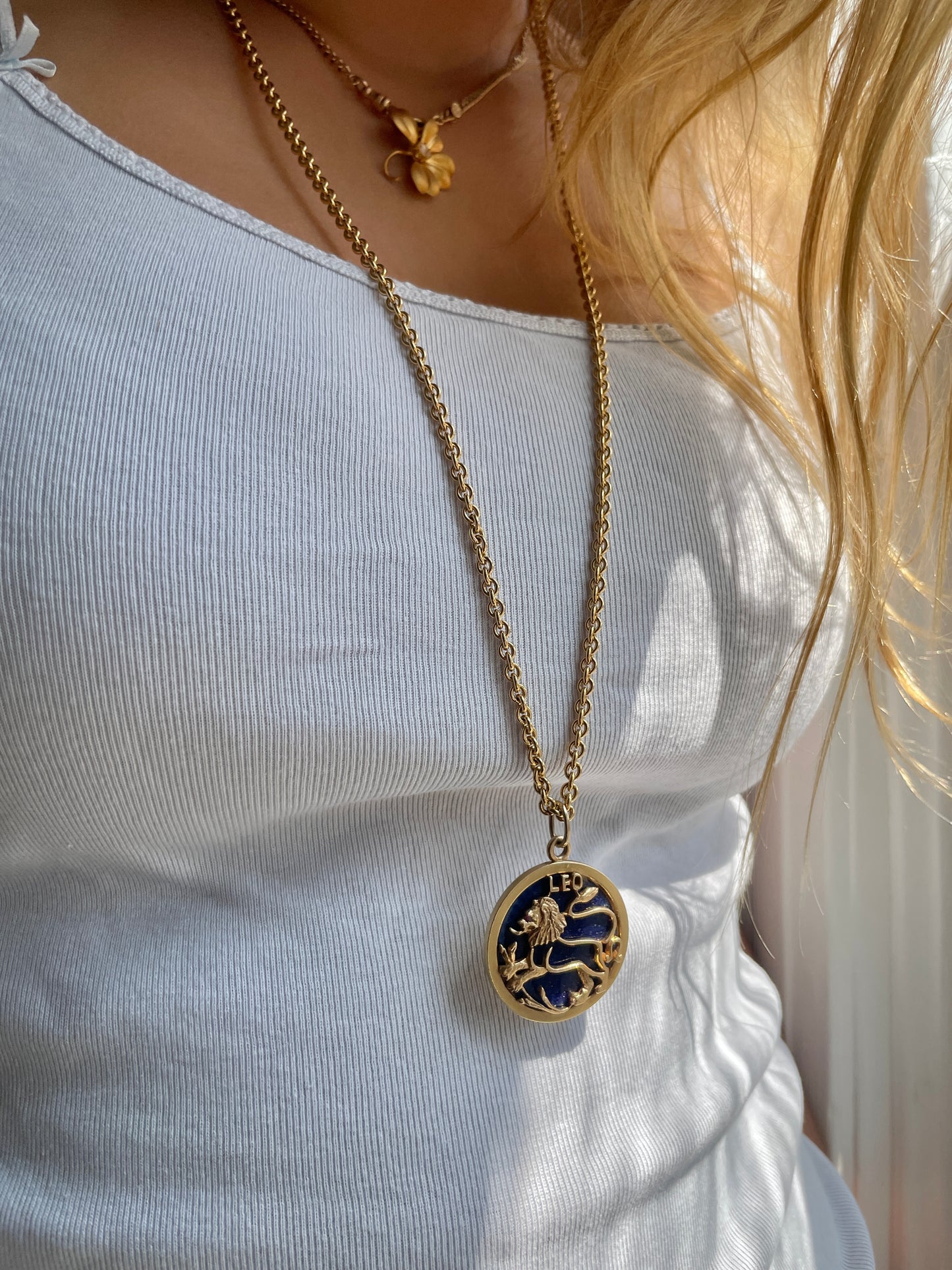 A gold  18 karat yellow necklace with a large round leo pendant worn by a person in a white top.